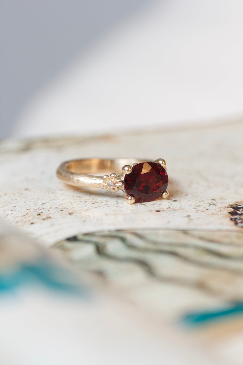 All about ring fra Pompus. Ringen er lavet i 14 karat guld og bærer en rød granat, samt to små brillanter fattet i hver deres blomst på siderne. Rustikt, men feminint udtryk. All about ringene skal male verden i farver og symboliserer alt, der giver værdi for den, der bærer den. Mens de emmer af farver og liv, gemmer de på et tomt lærred, der venter på sin historie og betydning. Hvad skal de symbolisere for dig? 